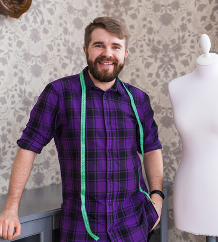 bhtpl A smiling man wearing a checked shirt with a measuring tape around his neck stands beside a mannequin.