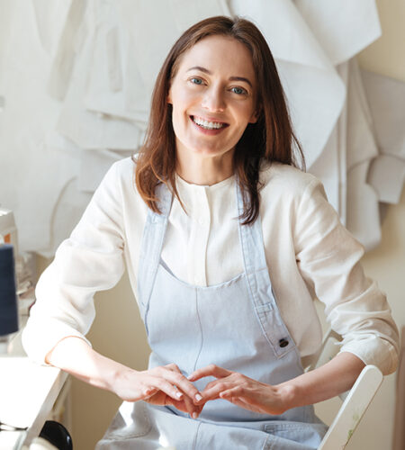 bhtpl A smiling woman wearing a white shirt and a light blue apron sits in an indoor setting with her hands gently clasped together on her lap.